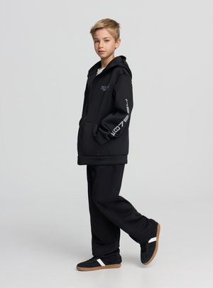 Ragazzo giovane in piedi con una gamba sollevata, indossa una felpa con cappuccio nera con testo bianco sulla manica, pantaloni neri e scarpe da ginnastica nere con striscia bianca.