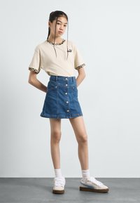 Kortermet beige t-skjorte med leopardmønster, kombinert med et blått denim A-linje skjørt med frontknapper og lommer. Hvite joggesko fullfører stilen.