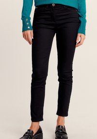 Jean leggings noirs à coupe slim, arborant un design à cinq poches et des détails matériels subtils. Assortis à un pull teal avec des accents de boutons.