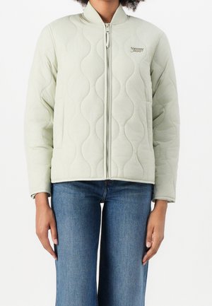 Kobieta ubrana w jasnozieloną pikowaną kurtkę na zamek z falistym przeszyciem oraz niebieskie dżinsy, stojąca na białym tle.