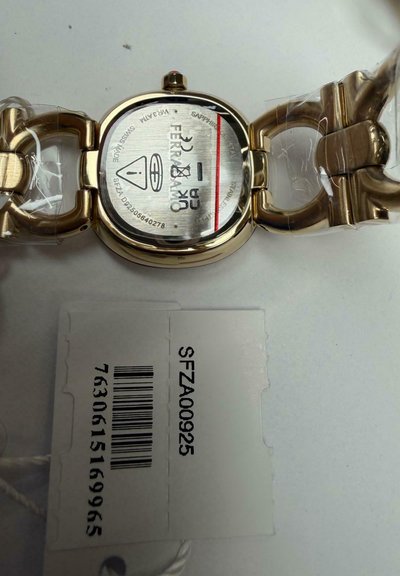 Horloge met goudkleurige wijzerplaat en ovale achterkant met merkaanduidingen, gravure in roestvrij staal en bevestigd wit label met barcode en productcode.