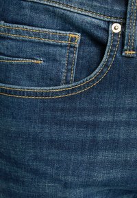 Tummat farkut, joissa on etutasku, kontrastivärillä kultaiset tikkauslangat ja sileä, teksturoitu pinta. Huomattava metallinen nappi.
