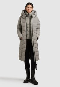 Langer grauer Daunenmantel mit Kapuze, der über gesteppte Abschnitte, einen Reißverschluss vorne und Seitentaschen verfügt. Getragen über einer ähnlichen grauen Innenschicht und schwarzen Leggings.