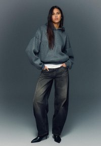 Veľká sivá mikina s textúrovaným povrchom, kombinovaná s voľnými čiernymi džínsami a špicatými čižmami na podpätku. Model stojí s rukami v rukávoch.