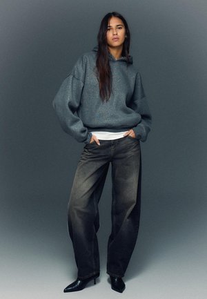 Übergroßer grauer Hoodie mit strukturierter Oberfläche, kombiniert mit lockeren schwarzen Jeans und spitzen Stiefeln mit Absatz. Das Modell steht mit den Händen in den Taschen.