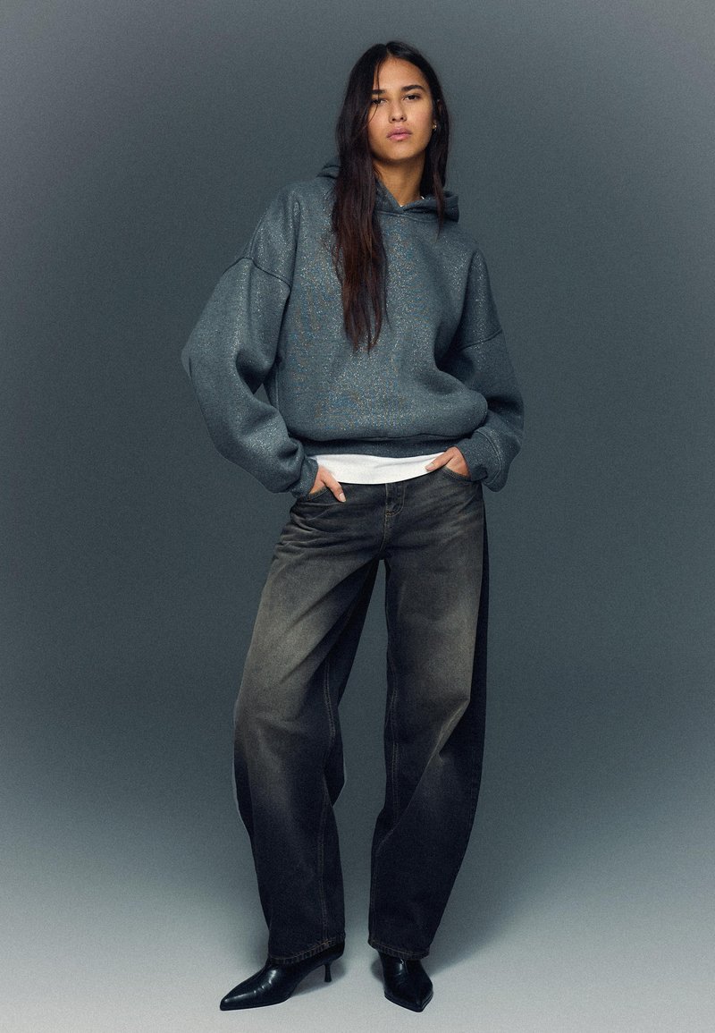 Veľká sivá mikina s textúrovaným povrchom, kombinovaná s voľnými čiernymi džínsami a špicatými čižmami na podpätku. Model stojí s rukami v rukávoch.
