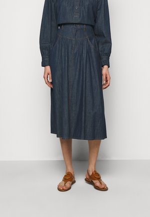 Vrouw draagt een donkerblauwe midi-rok van denim met zakken, een gerimpelde taille en een bijpassende denim top met lange mouwen en ceintuur, gecombineerd met bruine sandalen.