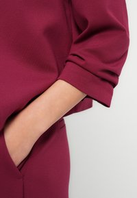 Πουκάμισο από υφασμάτινο ύφασμα σε μπουρντό απόχρωση με υφή, με μανσέτες γυρισμένες προς τα πάνω, διαθέτει πλαϊνή τσέπη με περιγραμμές από ομαλές ραφές. Απλός, σύ γχρονος σχεδιασμός.