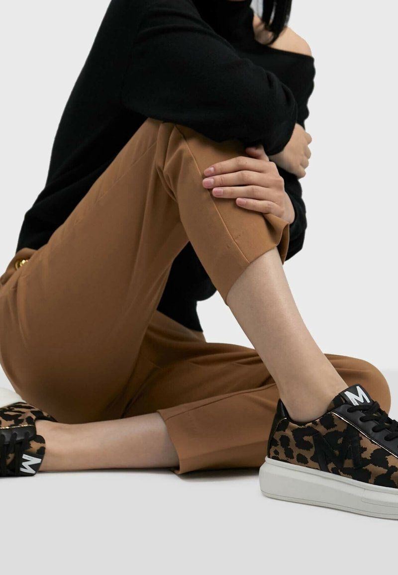 Sneaker nere e con stampa leopardo con un audace 'M' bianco sulla linguetta, indossate con pantaloni al polpaccio marroni. Texture liscia, design moderno e casual.