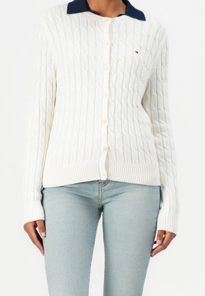 Žena v bílem zapínacím kardiganu s copánkovým vzorem a námořnicky modrým límcem, v světle modrých džínách, zobrazená od krku po boky.