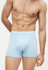 Torso masculin portant un boxer Calvin Klein bleu clair, montrant des abdominaux toniques et une partie des bras sur un fond uni.
