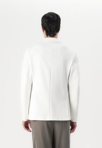 Blazer bianco con un design strutturato, una fessura posteriore singola e maniche lunghe. Realizzato in un tessuto morbido, abbinato a pantaloni grigi chiari.