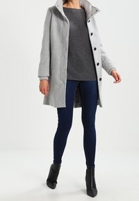 Abrigo gris claro con un cuello alto, botones negros y bolsillos laterales, llevado sobre un suéter gris oscuro y unos vaqueros ajustados azul oscuro, combinado con botines negros.