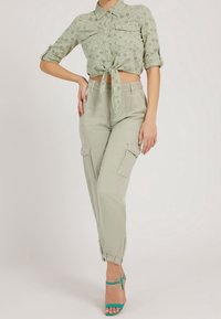 Woman wearing light green eyelet cropped shirt tied at waist, beige cargo pants with gathered ankles, and teal strappy high heels.