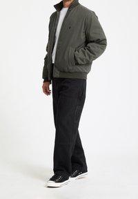 Homme portant une veste bomber vert olive, une chemise blanche, un pantalon cargo noir et des baskets noires, debout avec une main dans la poche.