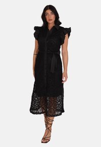 Abito in pizzo nero con maniche arricciate, chiusura con bottoni, vita cinturata e motivi floreali; orlo inferiore semi-trasparente. Sandali neri con strisce.