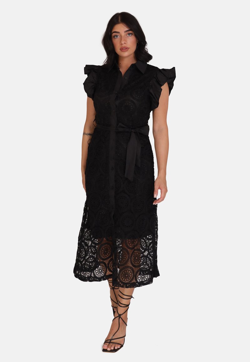 Abito in pizzo nero con maniche arricciate, chiusura con bottoni, vita cinturata e motivi floreali; orlo inferiore semi-trasparente. Sandali neri con strisce.