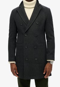 Dunkelgrauer, zweireihiger Wollmantel mit strukturierter Oberfläche, schwarzen Knöpfen und zwei Fronttaschen, kombiniert mit einem cremefarbenen Rollkragenpullover.