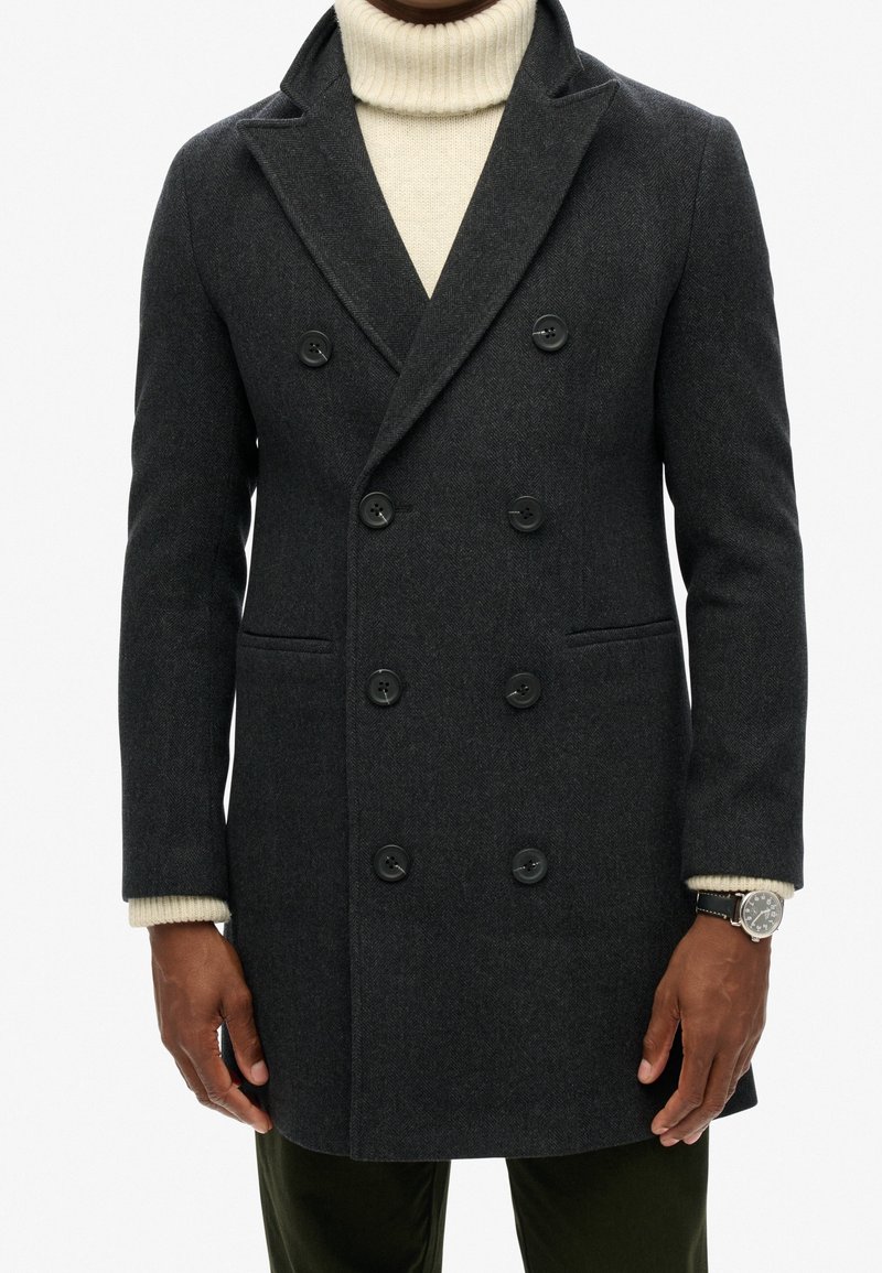 Dunkelgrauer, zweireihiger Wollmantel mit strukturierter Oberfläche, schwarzen Knöpfen und zwei Fronttaschen, kombiniert mit einem cremefarbenen Rollkragenpullover.