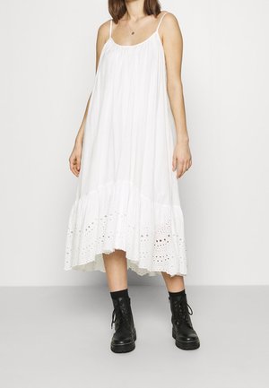 Femme portant une robe blanche ample sans manches avec un ourlet en dentelle ajourée, associée à des bottines noires à lacets et des chaussettes noires.
