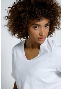 Bílé tričko s výstřihem do V a krátkým rukávem, měkká textura, hladký drapérií látky a minimalistický design, předváděné na modelce.