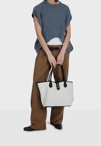Borsa a tracolla in tela con manici e dettagli in pelle nera, corpo di colore beige chiaro e dettagli stampati. Maglione grigio lavorato a maglia e pantaloni marroni.