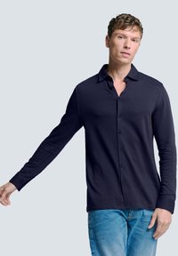 Marineblauwe overhemd met lange mouwen en kraag, knoopsluiting aan de voorkant, slim fit, zachte textuur, gecombineerd met lichtblauwe jeans, model poseert met één arm gestrekt.