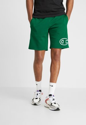 Mężczyzna w zielonych szortach Champion z dużym logo na lewej nogawce, czarnej koszulce, białych skarpetach Nike i butach sportowych stoi na szarej podłodze.