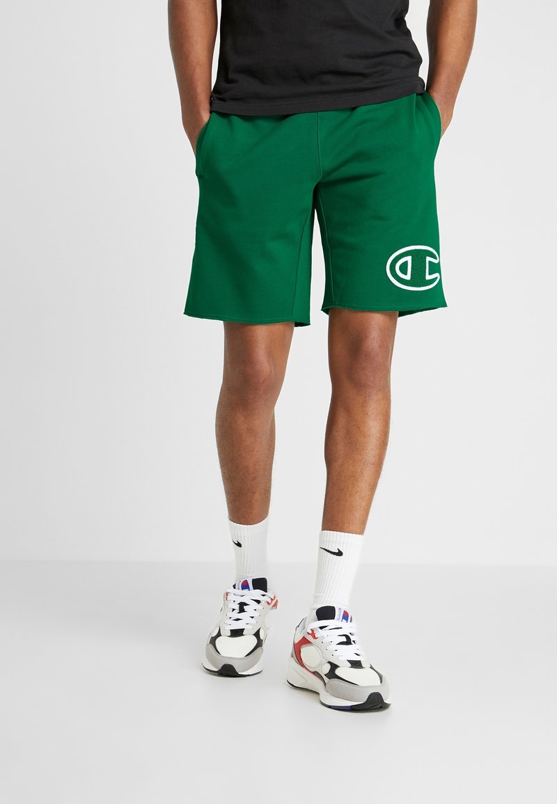 Homme portant un short vert Champion avec un grand logo sur la jambe gauche, un t-shirt noir, des chaussettes blanches Nike et des baskets, debout sur un sol gris.