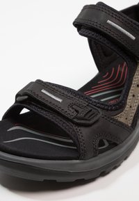 Musta ja harmaa sandaali, jossa on kaksi säädettävää Velcro-hihnaa, pehmustettu pohja ja tekokumin ulkopohja, jossa on punaisia aaltoyksityiskohtia.