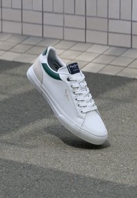 Baltās kedas ar teksturētu purngalu, zaļiem akcentiem uz apkakles un bežā zamšādas paneli uz papēža. Redzami šņores un logo uz mēles.