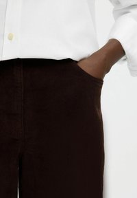 Ruka u džepu tamnosmešnih kord pantalona, nosi bijelu košulju dugih rukava s podignutim rukavima.