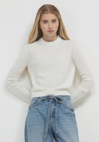 Junge Frau mit langen blonden Haaren, die einen weißen gestrickten Pullover und lockere blaue Jeans mit einem gebundenen Gürtel trägt, steht vor einer einfarbigen Wand.