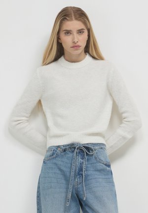 Giovane donna con lunghi capelli biondi, che indossa un maglione bianco lavorato a maglia e jeans blu larghi con una cintura annodata, in piedi contro un muro semplice.