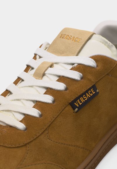 Baskets en daim marron avec lacets blancs, languette beige, et logo « VERSACE » visible sur la languette et le côté.
