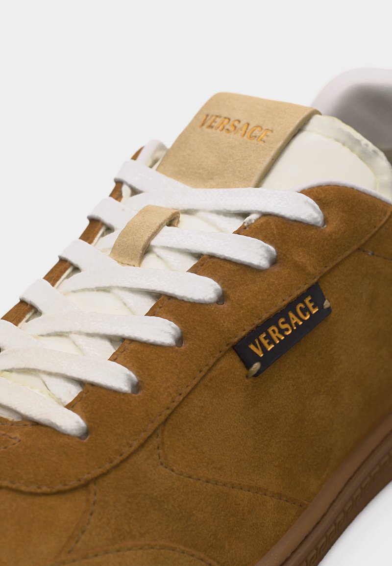Brauner Wildledersneaker mit weißen Schnürsenkeln, beigem Zungenstück und sichtbarem "VERSACE"-Logo auf Zunge und Seitenpaneel.
