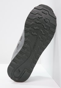 Chaussure de sport grise avec une semelle en caoutchouc texturé présentant un motif ondulé et un embout renforcé. Nom de la marque embossé sur le côté.