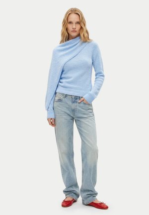 Frau, die einen hellblauen asymmetrischen Pullover, ausgewaschene weit geschnittene Jeans und rote Ballerinas trägt, steht vor einem unifarbenen hellen Hintergrund.