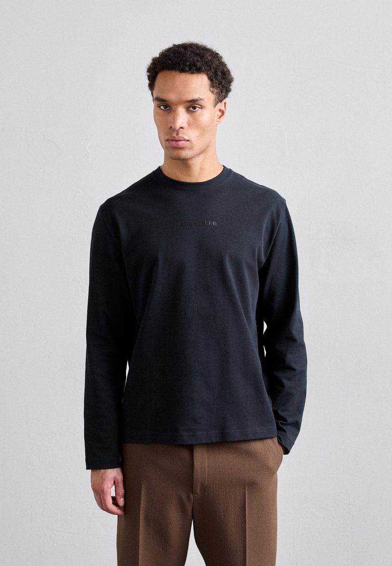 T-shirt noir à manches longues en coton doux, avec un col rond et un détail logo subtil sur la poitrine. Associé à un pantalon marron.