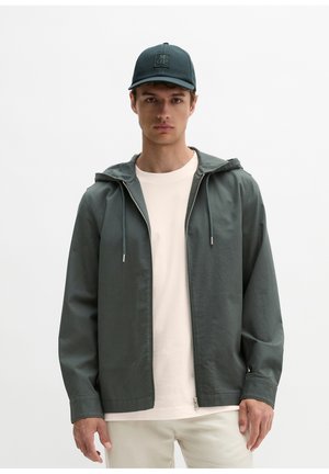 Mann trägt eine dunkelgrüne Kapuzenjacke mit Reißverschluss, weißes T-Shirt, beige Hose und eine dunkle Kappe mit quadratischem Logo vor weißem Hintergrund.
