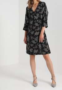 Schwarzes Kleid mit Blumenmuster, V-Ausschnitt, Dreiviertelärmel und taillierter Taille. Model trägt graue Riemchenheels.