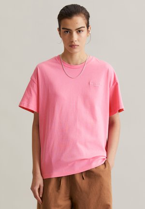 Jeune personne portant un t-shirt ample rose avec le logo "Marc O'Polo Denim" et un pantalon marron, debout devant un fond clair uni.