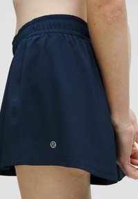 Marina atletiska shorts med elastisk midja, slät textur och en liten logotyp vid nederkanten. Kort längd med ett flödande design.