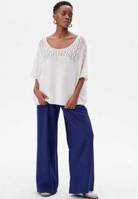 Lichtgewicht, wit gebreid shirt met een ruime halslijn en opengewerkte patronen, gecombineerd met losse marineblauwe broek en grijze sneakers.