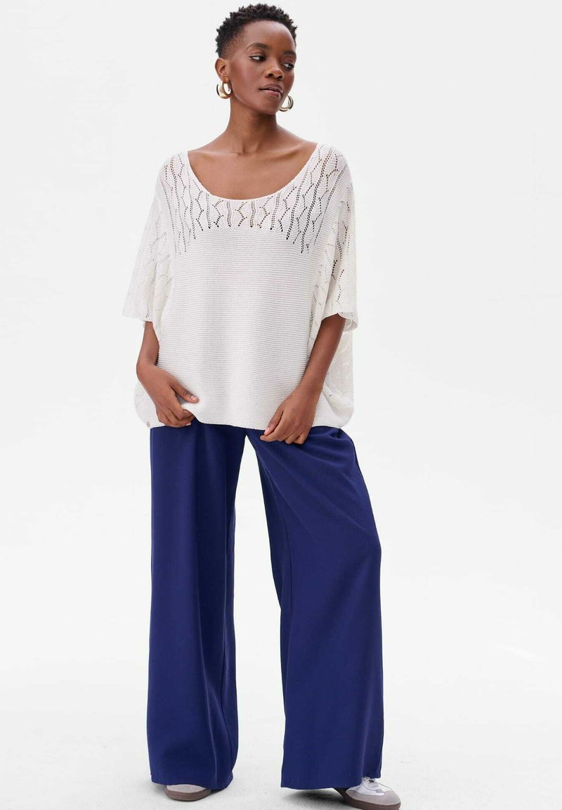 Lichtgewicht, wit gebreid shirt met een ruime halslijn en opengewerkte patronen, gecombineerd met losse marineblauwe broek en grijze sneakers.