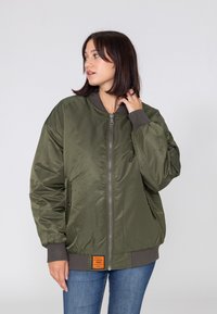 Veste bomber vert olive avec fermeture éclair à l'avant, poignets et ourlet côtelés. Présente un patch logo sur le devant. Tissu lisse et brillant, silhouette ample.