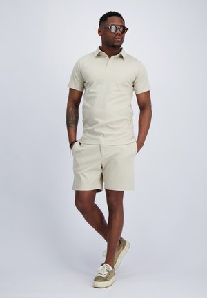 Mann trägt beiges Poloshirt, passende Shorts, Sonnenbrille und lässige Sneaker, steht mit den Händen in den Taschen vor einfarbigem Hintergrund.