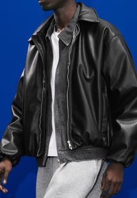 Črna usnjena bomberska jakna z teksturiranim zaključkom, ki ima visok ovratnik, sprednji zadrgo in elastične manšete. Nosi se čez belo srajco.