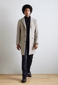 Abrigo de lana gris con cuello notch, camiseta polo negra y pantalones a rayas. Zapatos negros con textura suave. Modelo de pie.
