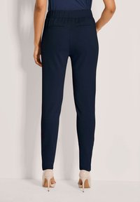 Vrouw in marineblauwe tapered broek en beige hoge hakken staat tegen een lichte muur, van achteren gezien.